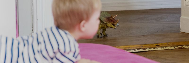 Ein Kind liegt bäuchlings auf dem Boden. Neben ihm steht eine Dinosaurier Spielfigur. Ein Triceratops.