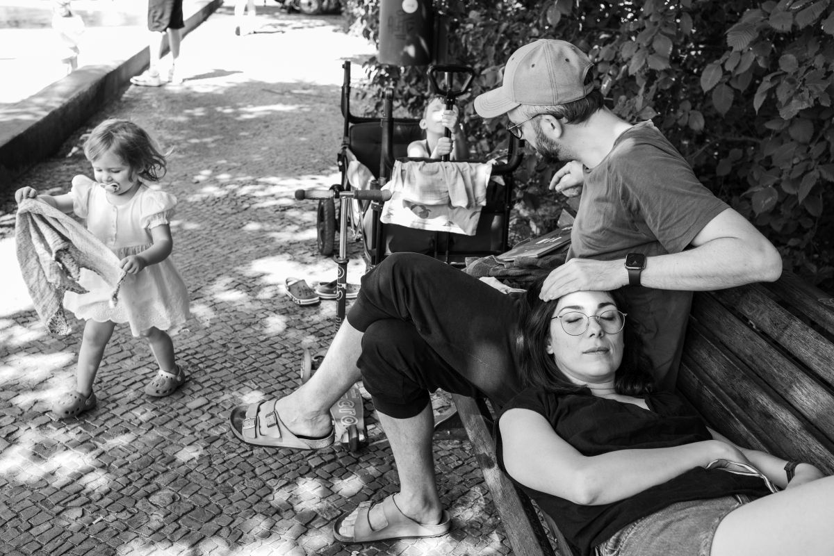 Eltern auf einer Parkbank. Die Mutter hat ihren Kopf auf den Schoß des Vaters gelegt und die Augen geschlossen. Zwei Kinder spielen drum herum.