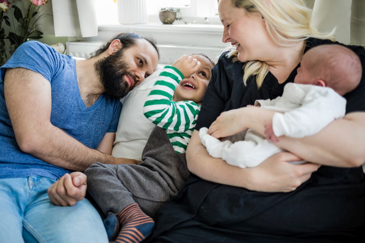 Eine Mutter hält ihr Neugeborenes und lächelt dabei ihr Kleinkind an. Der Vater beobachtet dies lächelnd.