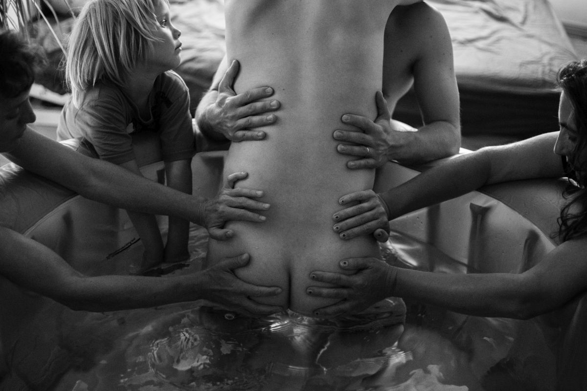 Eine Frau wird im Gebärpool von ihrem Mann gehalten und zwei Hebammen stützen ihr Becken während einer Wehe. Nebenbei spielt ein Kind mit seinen Händen im Wasser.