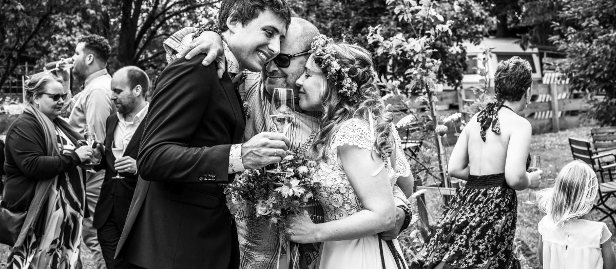 Ein Hochzeitspaar wird liebevoll von einem Familienmitglied umarmt. Im Hintergrund feiern andere Gäste.