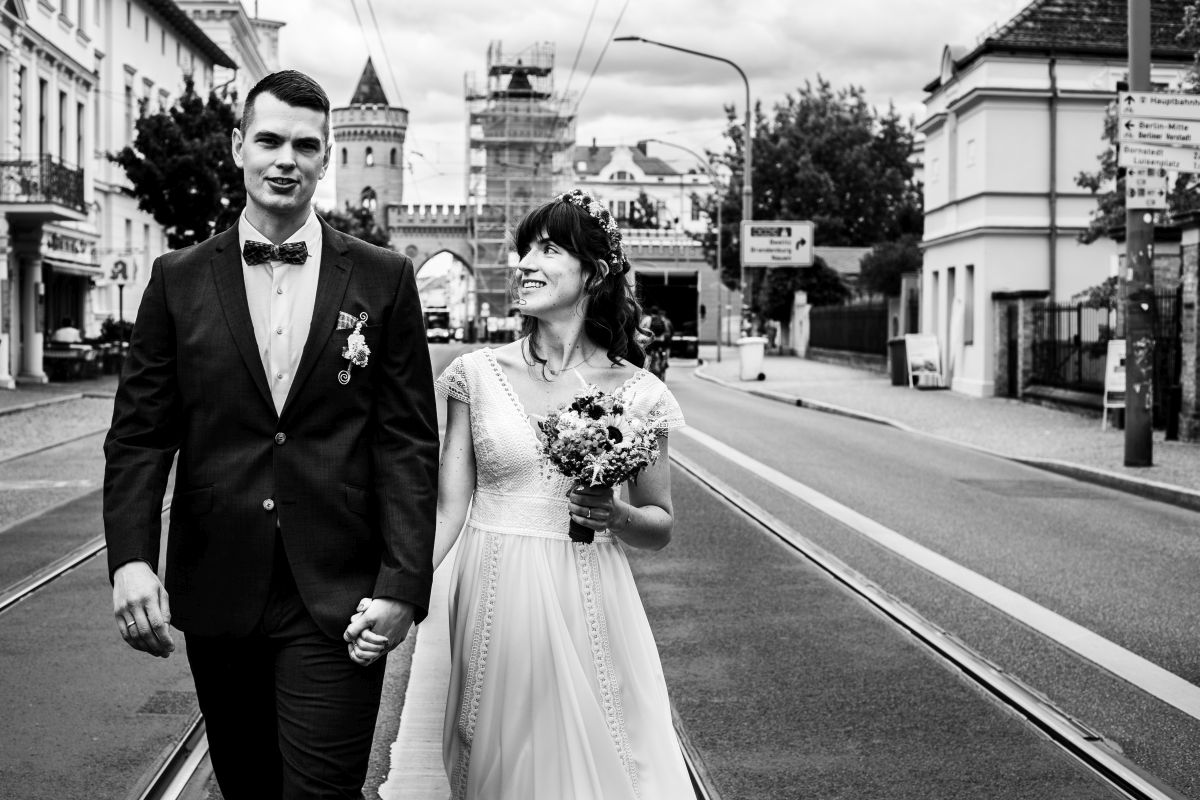 Ein Hochzeitspaar vor dem Nauener Tor in Potsdam. Die Braut schaut den Bräutigam an während sie Hand in Hand auf der Fahrbahn entlanggehen.