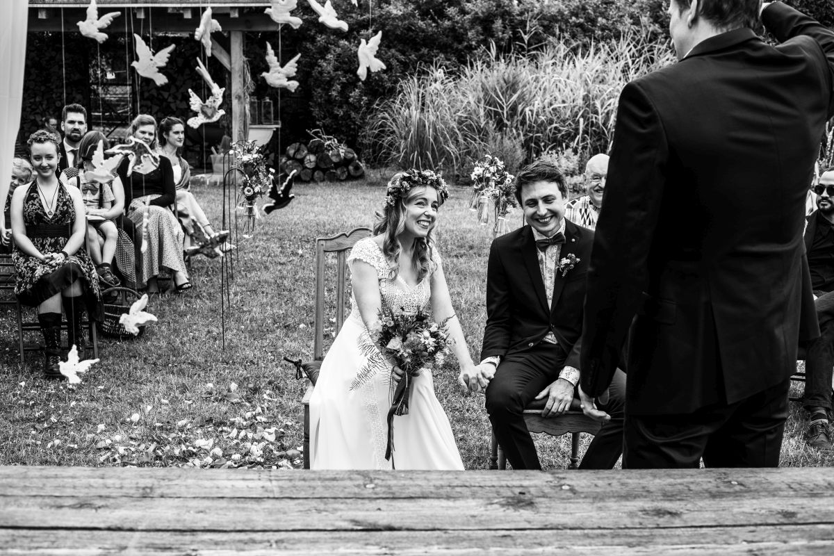 Ein Brautpaar sitzt lachend vor dem freien Trauredner. Sie halten sich an der Hand. Rechts von ihnen hängen kleine Tontauben als Deko. Hinter ihnen sitzen die Hochzeitsgäste.